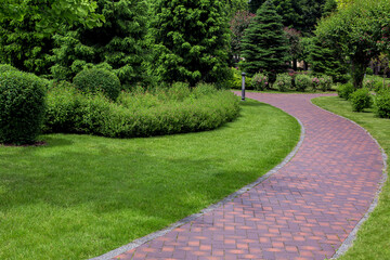 curved pathway paved brick stone tiles in park among plants, foliage bushes and pine trees...