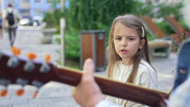 Portrait of serious pretty little girl singing to the accompaniment of a guitar outdoor. Cute child girl with long blonde hair singing sitting on the bench on the street. High quality 4K footage