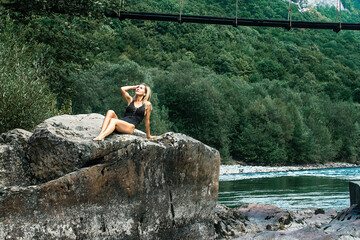 a beautiful slender blonde girl in a dark joint swimsuit sits on a large stone near a mountain river. Rest in exotic places of nature