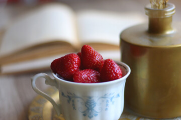 Vintage cup with fresh strawberries, brass tray, vase with gypsophila flowers and open books on the table. Selective focus.