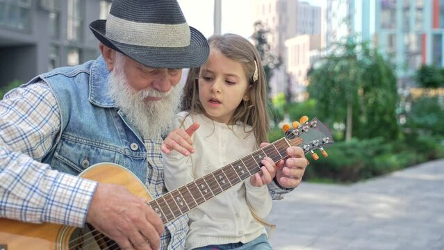 Zoom out caring senior caucasian grandpa teaching his lovely enthusiastic little granddaughter to play guitar sitting on a bench outdoor on the street. High quality 4K footage 