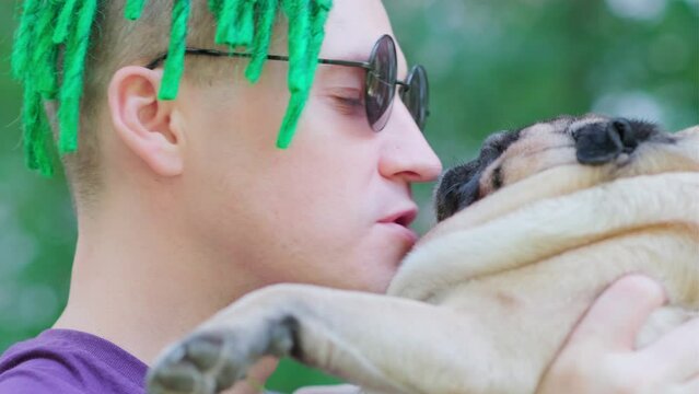 Portrait of man with green dreadlocks in sunglasses holding beige pug in hands, kissing dog on muzzle. Close up.