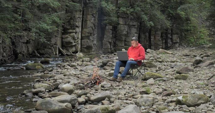 A man works on a laptop near a stream in nature