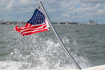 American flag flapping in the wind at the stern of a boat, showing water splashing and coastline in the background.