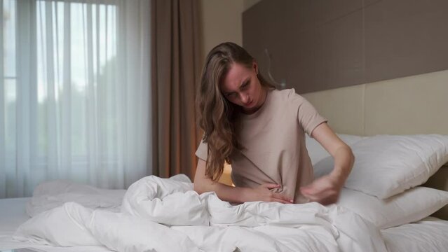 Woman curls up touching stomach with hands sitting on bed in hotel room. Sick female person feels pain in abdomen after waking up in early morning