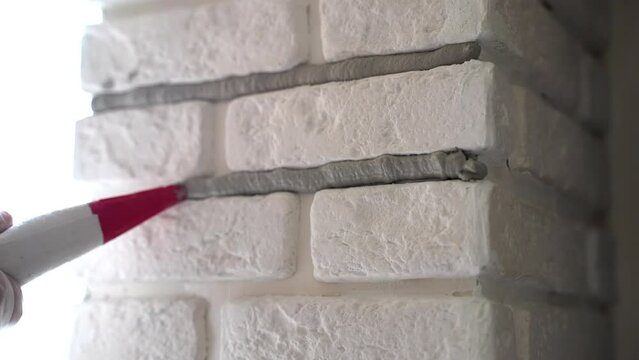 Workman in protective gloves filling joints with grout using construction syringe, builder finishing white decorative stones on interior wall. Applying mortar between stone plates, wall restoration