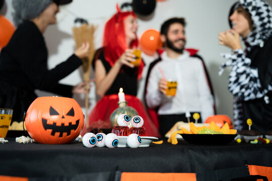 Group of friends in a halloween party. Devil, witch, dracula and harlequin eating, drinking and laughing