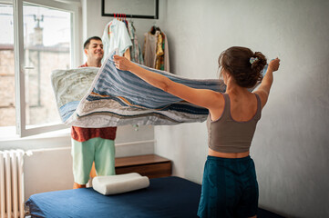 Cheerful young couple making bed, having fun at home. Doing house chores together, cleaning up in...