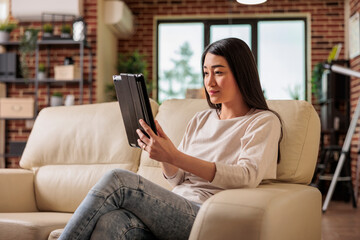 Obraz premium Beautiful young chinese woman using digital tablet at home, connecting internet web 3.0 smiling while sitting on couch entertainment device, browsing internet wireless device computer tablet using