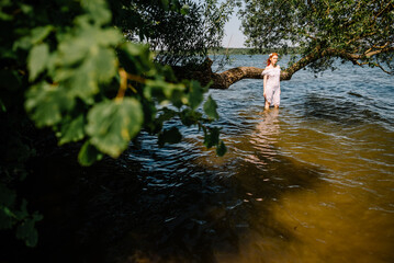 Woman in summer white dress stands on the seashore and looks at the horizon. Young beautiful girl standing in the water