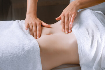 Obraz na płótnie Canvas Spa procedure in a massage room, a woman patient lies relaxed on the table covered with a white terry towel, gentle hands of a female massage therapist massage her belly