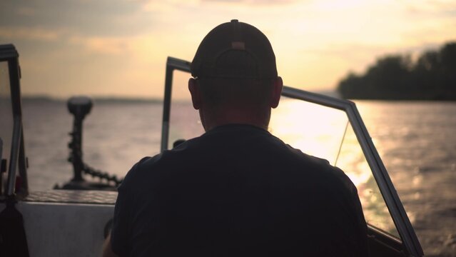 A man drives a fishing boat. A fisherman is sailing in a boat on the river at sunset. Back view of a man in a cap.
