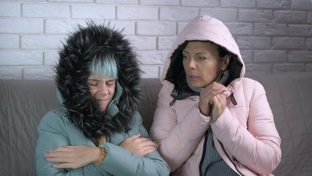 Family freeze in room. A freezing woman with her daughter in warm coats sits on the sofa in the room. A concept of unheated winter period during energy inflation.