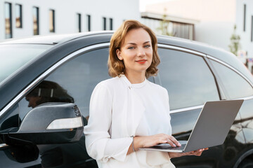 Portrait of confident smiling manager using laptop standing near new car. Dealership, car rental concept  