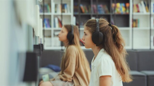 Girls sitting in class wearing headphones and looking at device on wall, focus on child in front, bookshelves on background. Arc shot pupils studying at school. Concept of education
