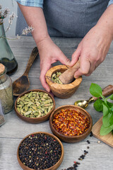 Woman's hands grind cardamom in a wooden mortar. Kitchen gray table with various spices.