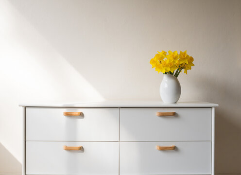 Yellow daffodils in white vase on side table against beige wall with light from window (selective focus)