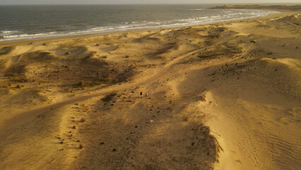 Couple with dog walking on sandy dunes of La Viuda beach at sunset with Punta del Diablo village in...