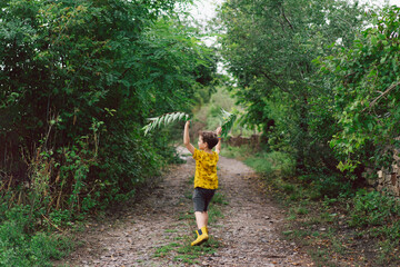 Naklejka na ściany i meble A six-year-old boy runs with green leaves in his hands in the countryside. Happy child boy laughing and playing in the summer day. Back view