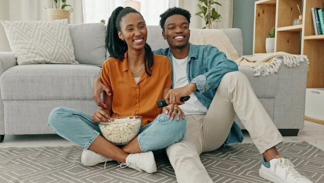 Happy black couple, eating popcorn and a movie playing in the living room of their home or house. Love, romance and man and woman having leisure time while looking at tv and browsing channels.