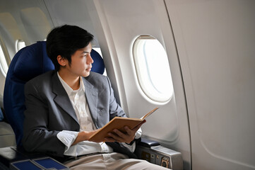 Successful Asian businessman holding a book while looking out the window, rduring the flight