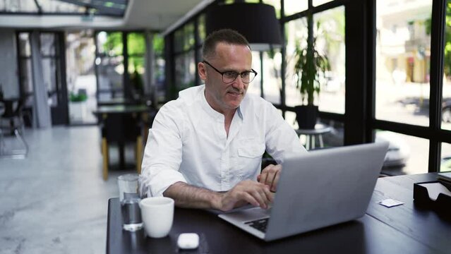 Smiling businessman working on laptop computer at caffee. Male professional typing on laptop keyboard at caffee workplace. Portrait of positive business man looking at laptop screen indoors