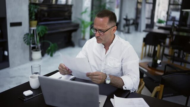 Focused adult businessman working on project, writing paper notes, sitting at caffee desk. Concentrated male professional analyzing market research information