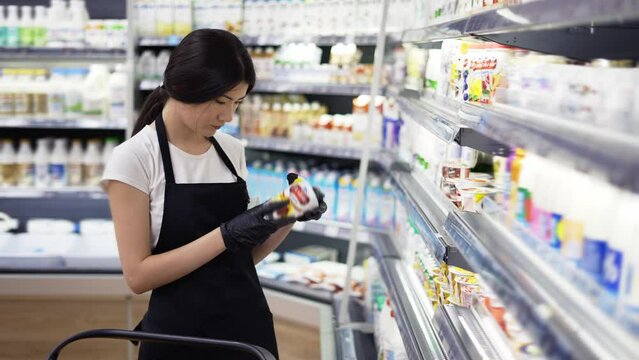 Asian Girl Worker In An Apron With A Badge Checks The Goods On The Shelves. Re-inventory Of Goods, Checking The Expiration Date Of Goods