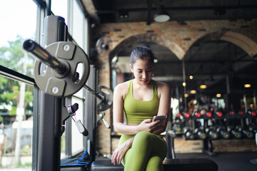 Beautiful young Asian woman in sportswear resting or waiting her friend after exercise at fitness gym and using smartphone for communication chatting or social media. Healthy lifestyle.