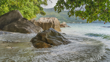 The waves of the turquoise ocean break on picturesque boulders and foam on the sand. The green...