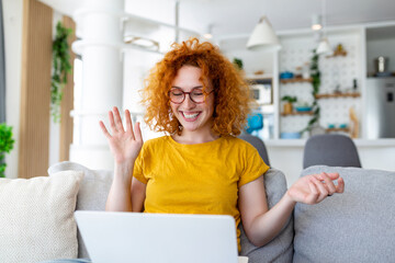 Happy young woman having fun doing video call using laptop in her home, waving hand video conference calling on laptop computer sit on sofa distance learn zoom online virtual meeting at home.