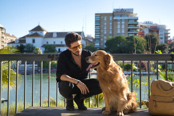 A young Hispanic man with a beard, sunglasses, black shirt and backpack, climbed on a bench with his dog while petting him under the sun's rays. Concept animals, dogs, love, pets, golden.