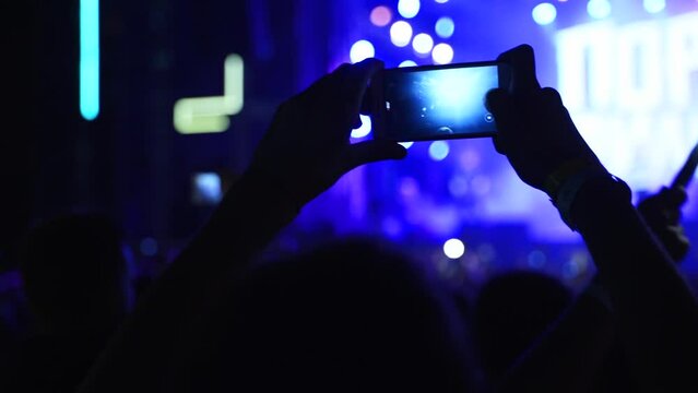 People silhouettes waving hands, taking photos or recording videos of live music concert with smartphones at open air festival. Bright colorful stage lighting. Fans at favourite band performance.