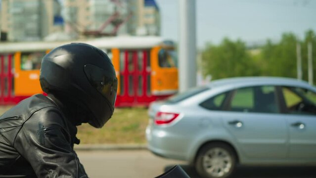 Motorcyclist man in black helmet with closed visor and leather jacket rides modern cycle along large city street on sunny day side view slow motion