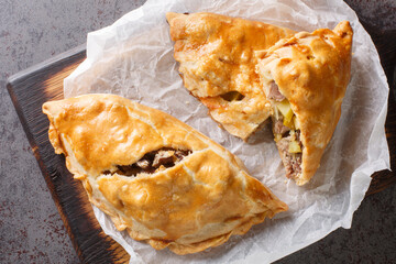 Traditional Cornish pasty filled with meat potato swede and carrot closeup on the paper on the wooden board. Horizontal top view from above - obrazy, fototapety, plakaty