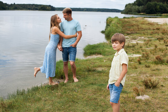 A Couple In Love Are Standing On The Beach, On The Banks Of A Picturesque River. Their Son Is Standing Next To Them. Mom Is Wearing A Blue Dress, Dad And Son Are Wearing Denim Shorts And T-shirts.