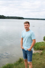 A man with an athletic body, dressed in denim shorts and a blue T-shirt, stands on the banks of a picturesque river during the day.