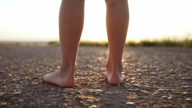 Close up of bare foot and nake legs of unrecognizable person who stands on asphalt road. Nature field and plants. Part body wandering person. Health care and meditation, therapy. Sunbeams in summer.