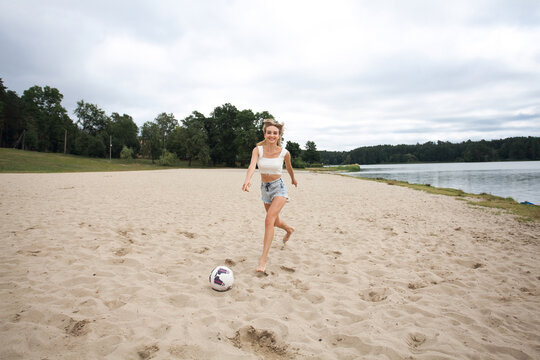 A beautiful slender girl with long blond hair and long legs runs along the beach towards a soccer ball, wearing short sexy denim shorts and a white tank top.