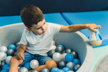 Toddler boy playing with a toy while sitting in a ball pit full of colorful balls. A ball pit - a great place for kids to jump, crash, and wiggle. Sensory play at the nursery school. . High quality - obrazy, fototapety, plakaty