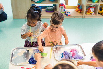 Sensory play at multi-cultural nursery school. Toddlers with their teacher playing with striped straws and milk painting, using food coloring, milk, watercolor paper, and trays. Creative kids activity