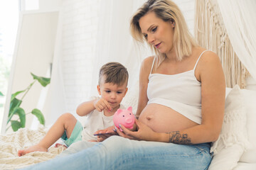 Finance responsibility from a young age. Indoor shot of two caucasian people - pregnant mother in her 30s wearing white tank top in a way that shows her pregnant belly and little toddler boy sitting