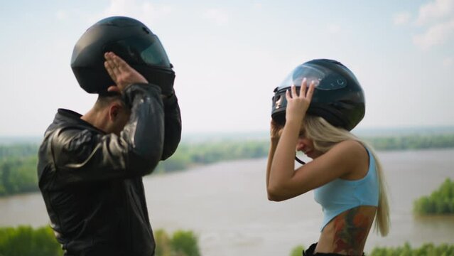 Happy man and young woman in blue top take off protective moto helmets standing against large picturesque lake on spring day slow motion