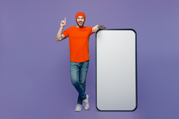 Full body insighted young man wear red hat t-shirt big huge blank screen mobile cell phone workspace mockup area hold index finger up great new idea isolated on plain pastel light purple background.