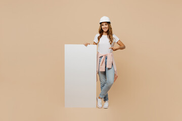 Full body young employee handyman woman in white t-shirt helmet show big white blank billboard wall isolated on plain beige background Instruments accessories for renovation room. Repair home concept.