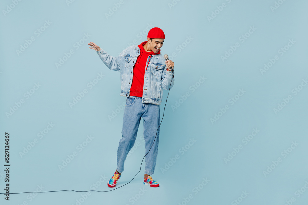 Wall mural Full body young singer middle eastern man 20s he wear denim jacket red hat sing song in microphone at karaoke club isolated on plain pastel light blue cyan background studio. People lifestyle concept.