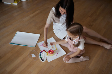 Smiling young mother and little daughter lie on a warm wooden floor in the living room, drawing together, happy mother rejoices at her daughter's drawings. paint on canvas