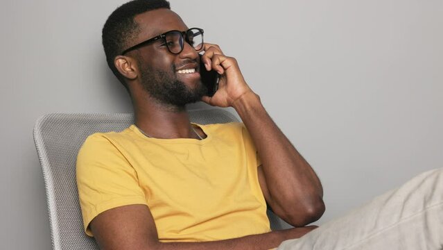 African-American man sitting in a chair at home in a yellow T-shirt with a phone talking with glasses and a smile with teeth close-up, technology for people