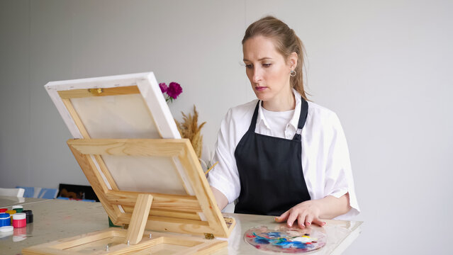 Mature woman paints picture on easel sitting at table. Professional artist enjoys painting masterpieces with paintbrush and palette closeup