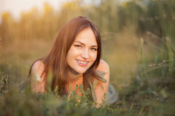 Naklejka na ściany i meble Brown-haired woman lies on grass admiring picturesque countryside landscape. Lady looks around with excited and smiling expression on meadow, sunlight